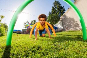 Young boy crawling on the grass through colorful pool noodle arches in an outdoor obstacle course on a sunny day.