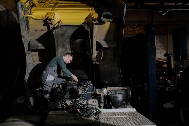 A mechanic works on a truck engine with the cab lifted, performing maintenance inside a dimly lit workshop.